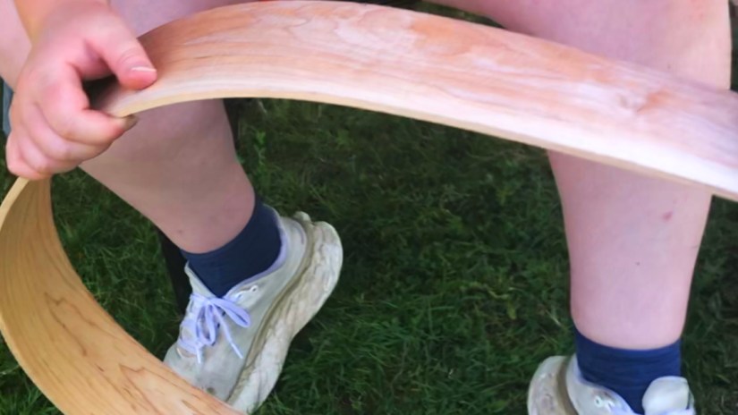 Hands bending a wooden drum frame over the knees of a person seated on grass.