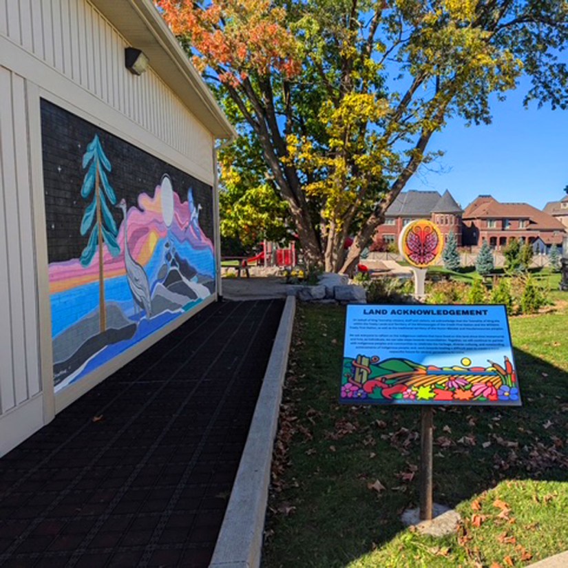 A municipal Land Acknowledgement sign featuring Portia “Po” Chapman’s Storytelling Art illustration, installed in a King Township garden alongside an Indigenous mural and butterfly art sculpture.