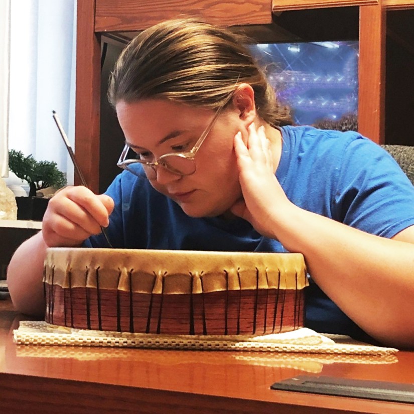 Kingston Artist Portia “Po” Chapman in her studio, meticulously hand-painting a custom rawhide frame drum for a collector using a small round artist brush.