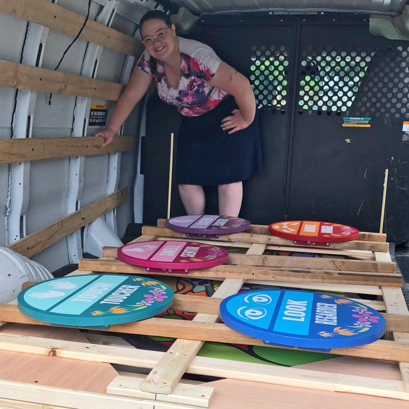 Kingston artist Portia "Po" Chapman posing inside a delivery van with her custom-built layered crate securely holding the Sensory Garden mural and five circular station signs.