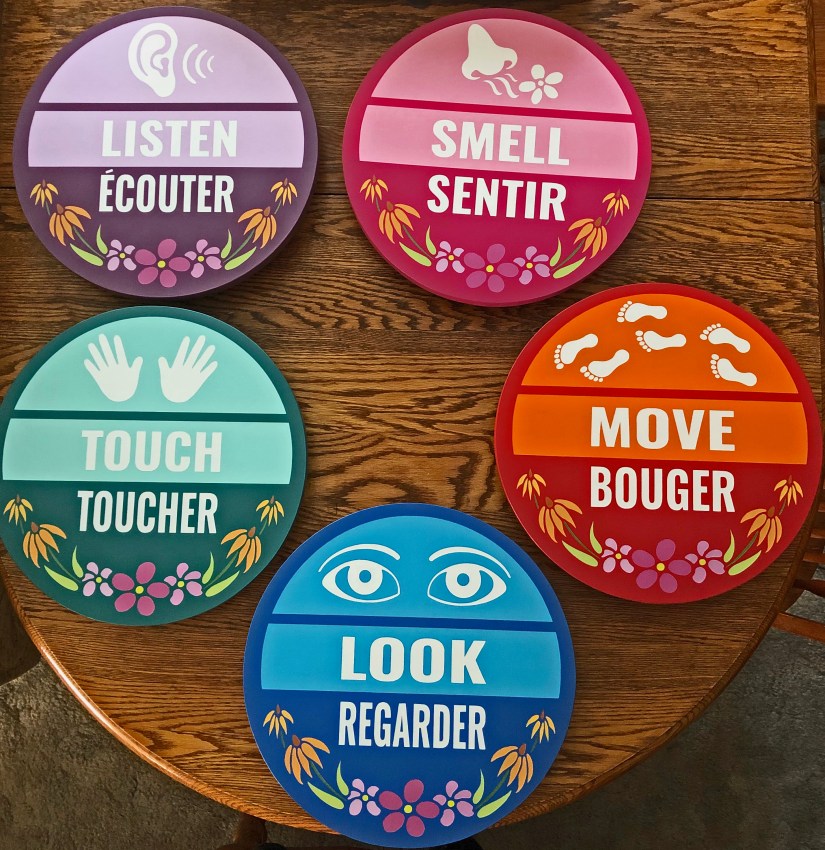 Five circular, hand-painted station signs for the Sensory Garden—Listen, Smell, Move, Touch, and Look—presented in their final bilingual format on a wooden table.