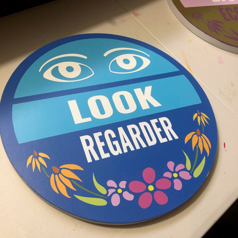 A close-up of the finished "LOOK / REGARDER" circular sign for the Sensory Garden, showing the hand-painted bilingual text, eye icons, and vibrant floral motifs.