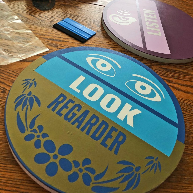 A finished circular sensory garden sign on a wooden table, featuring the words "LOOK" and "REGARDER," eye icons, and floral motifs that match the main mural.