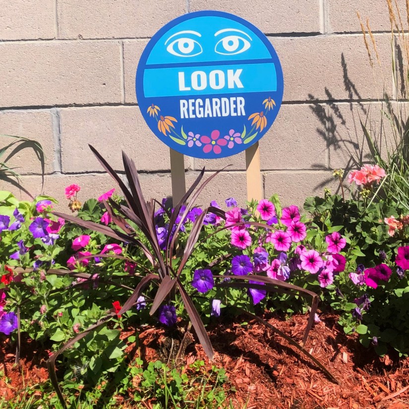 The finished "LOOK / REGARDER" bilingual circular sign standing in a studio flower garden to test visibility and colour harmony before final installation.