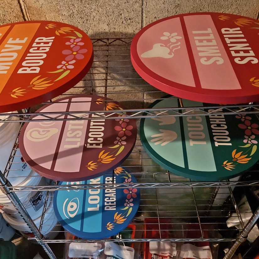 Five circular bilingual station signs for the Sensory Garden—including "LOOK/REGARDER" — drying on a wire rack after receiving a protective UV-resistant top coat.