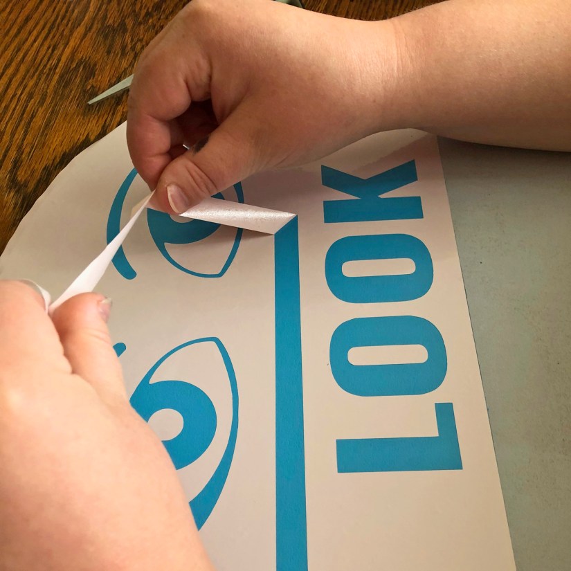 Close-up of Portia "Po" Chapman peeling CNC-cut vinyl elements from a custom "LOOK" station sign for the Sensory Garden project.
