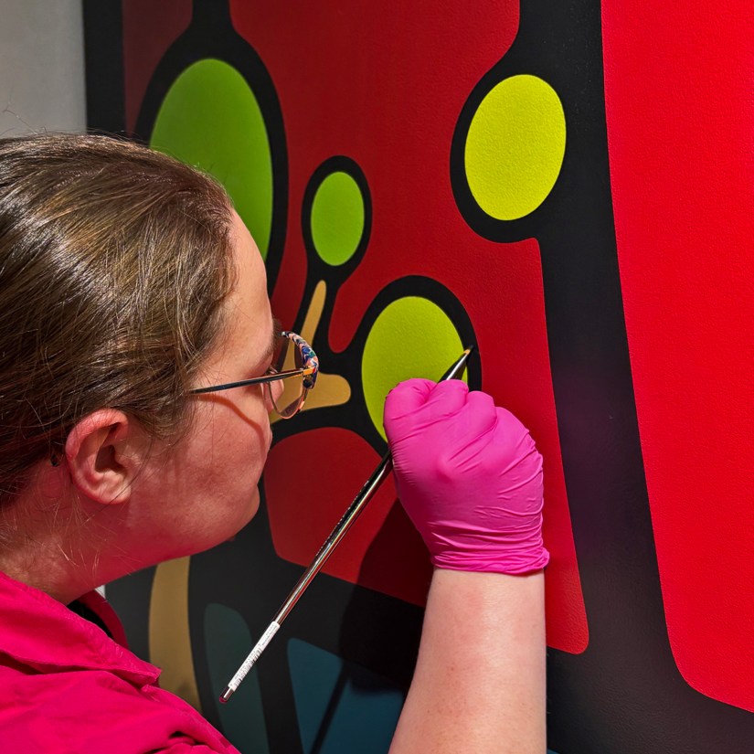 A close-up of Kingston Artist Portia “Po” Chapman hand-painting the "Strong and Growing Stronger" mural with a small flat brush, showcasing uniform texture, deep color saturation, and crisp line-work.