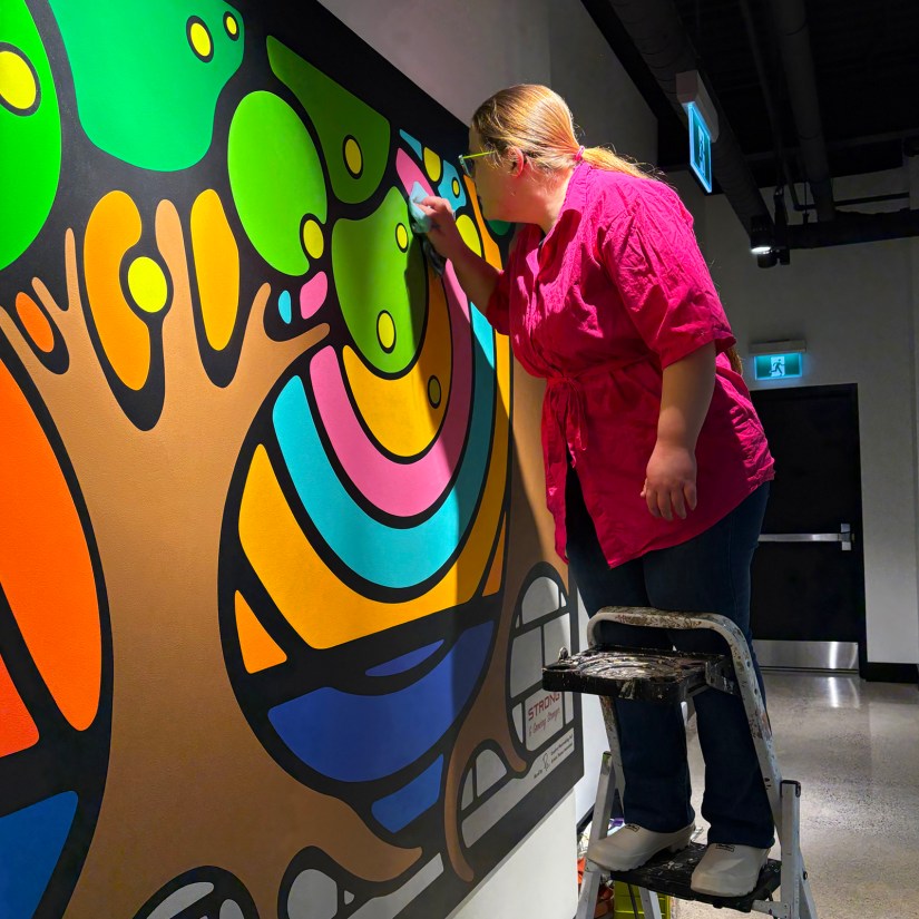 Kingston Artist Portia “Po” Chapman on a ladder polishing her "Strong and Growing Stronger" mural at night, showcasing crisp free-hand line-work and unique, hand-mixed colour blocks in a professional office installation.