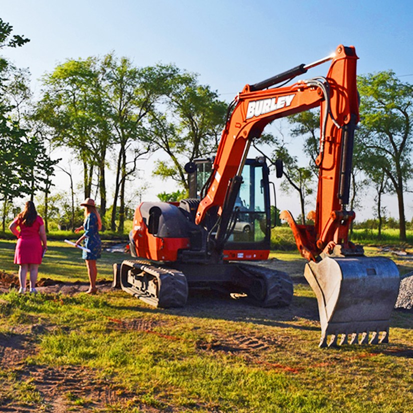 Installation Artist Portia Chapman in a blue dress and hat, holding architectural plans, standing in a field with Wild Child Regeneration CEO Jessica Pelchat and a large orange excavator.