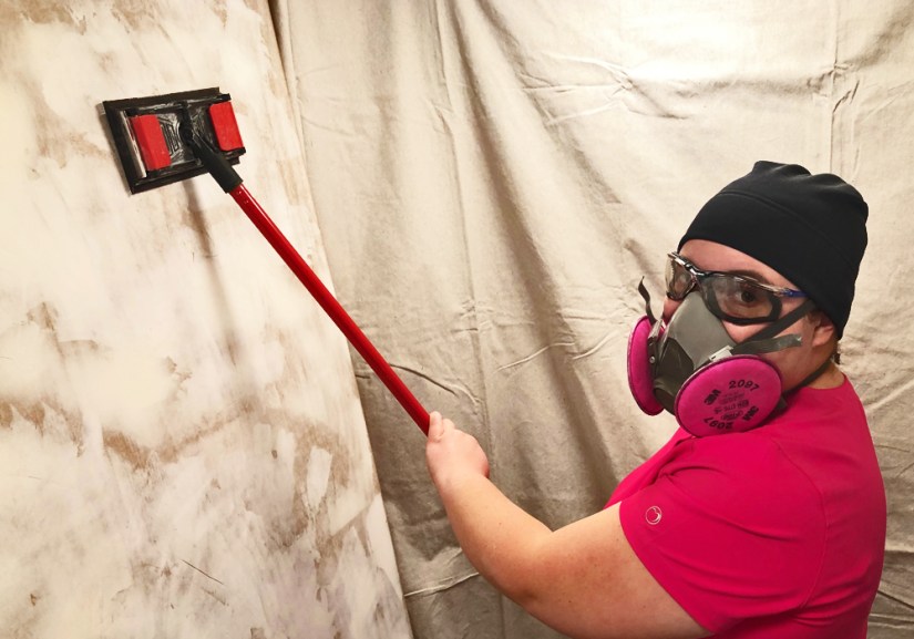 Kingston Artist Portia Chapman wearing a respirator and safety gear while using a pole sander to smooth the filled surface of a large 14-foot mural board.