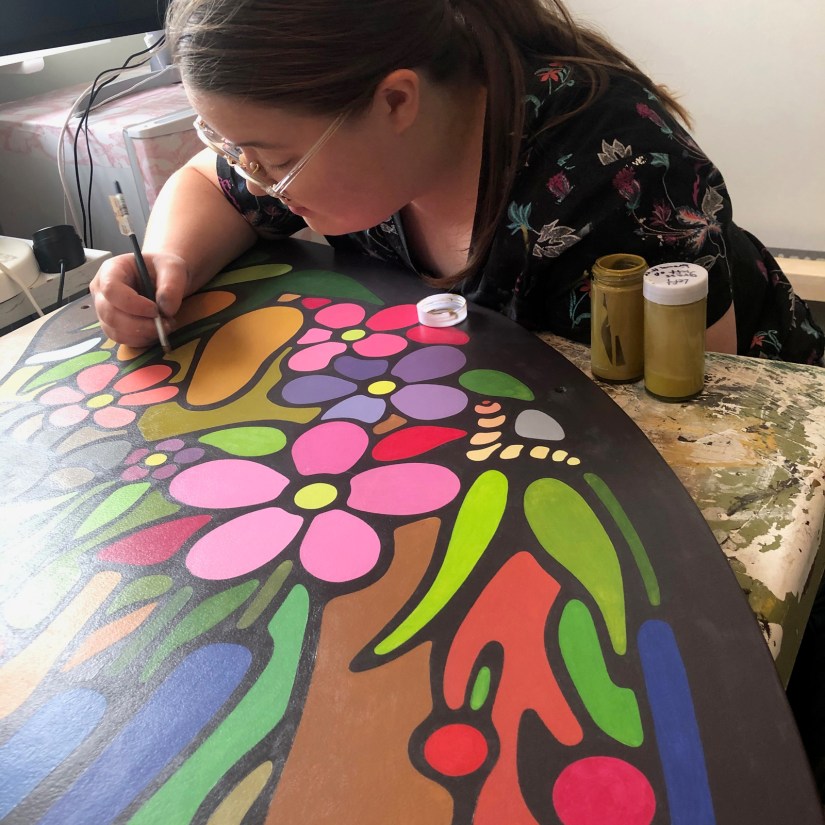 Kingston artist Portia "Po" Chapman using a small round artist brush to apply a precise second coat of paint and black line-work to the Sensory Garden mural.