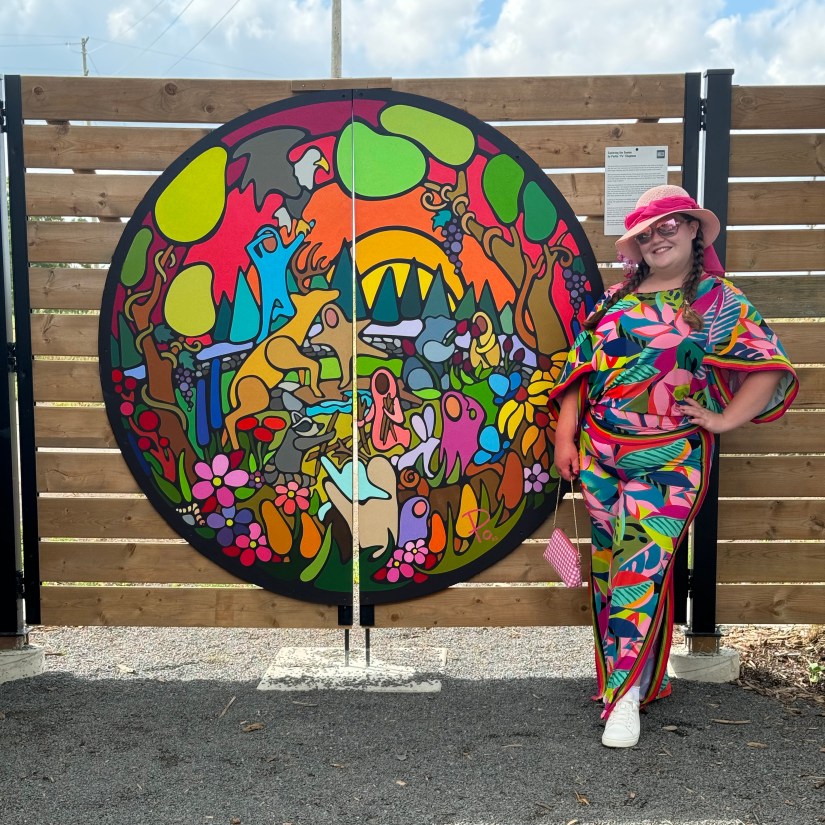 Kingston artist Portia "Po" Chapman standing beside the installed "Exploring the Senses" Storytelling Art mural on the Sensory Garden gate at Base31.