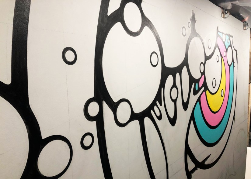 A close-up process shot of the "Strong and Growing Stronger" mural showing the first application of yellow, pink, and teal paint within black-brushed outlines.