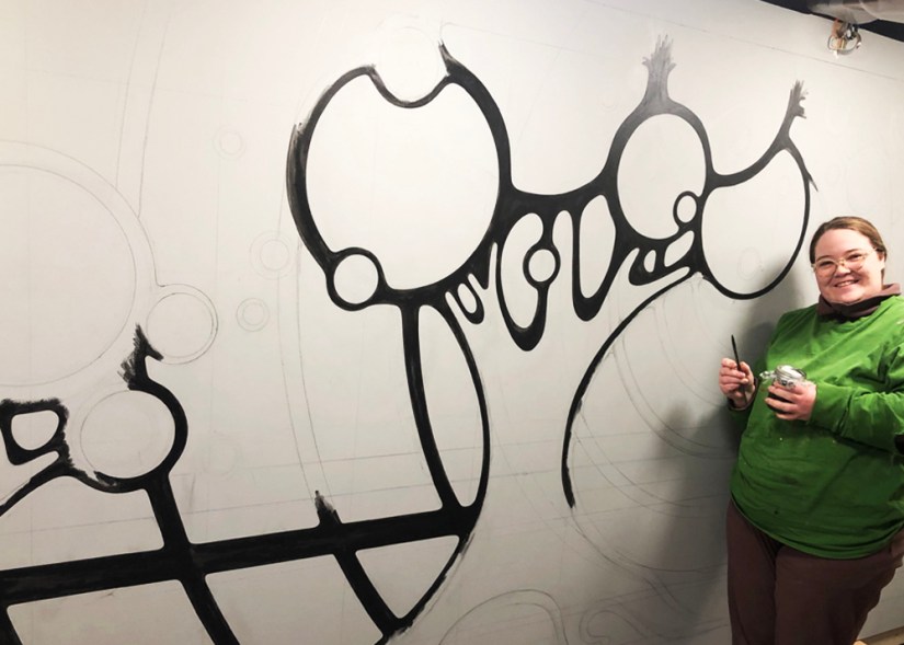 Kingston Artist Portia Chapman hand-painting black line-work on the "Strong and Growing Stronger" mural, following freehand pencil-drawn guidelines on a large-scale surface.