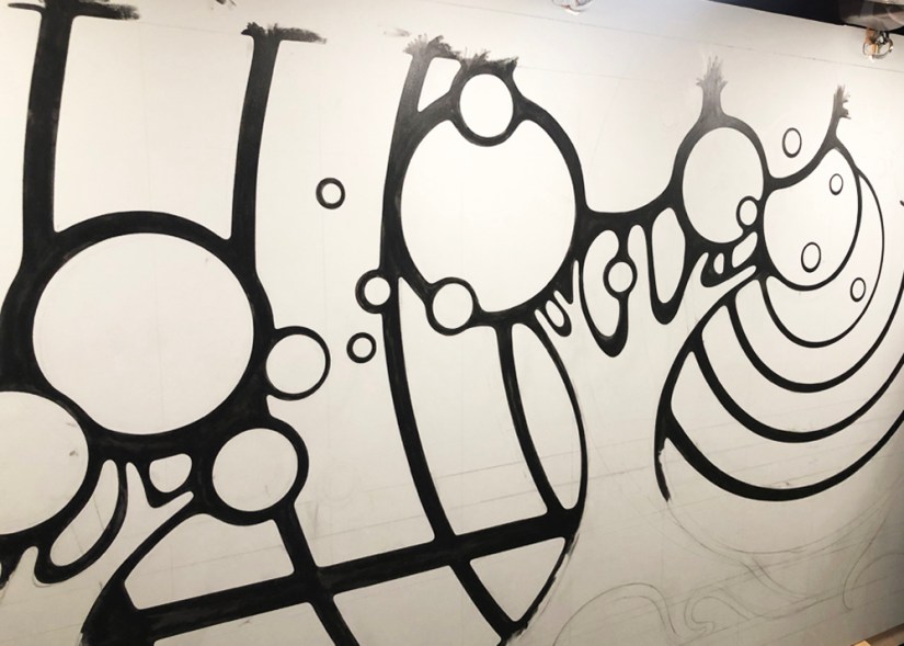 Kingston Artist Portia Chapman hand-painting black line-work on the "Strong and Growing Stronger" mural, following freehand pencil-drawn guidelines on a large-scale surface.