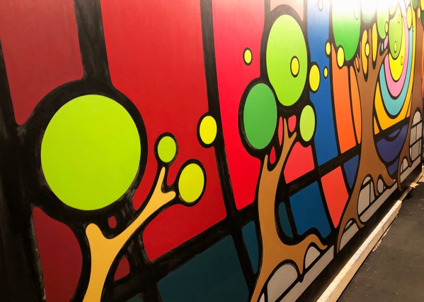 A close-up process shot of the "Strong and Growing Stronger" mural showing vibrant color blocks after three coats of hand-brushed paint, featuring crisp transitions as saturation builds with each additional coat.