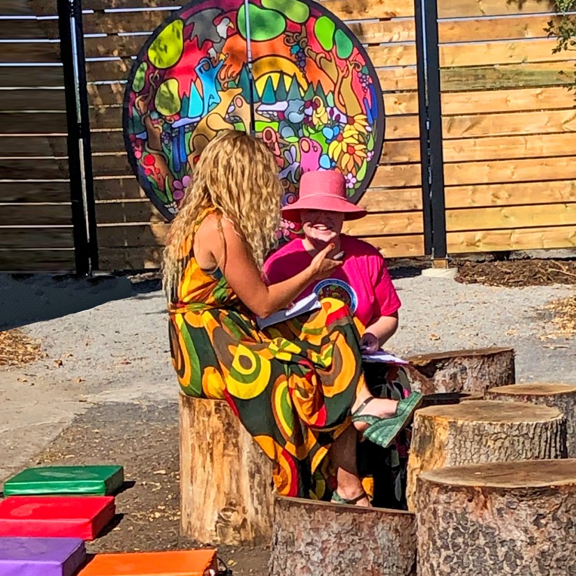 Kingston Artist Portia "Po" Chapman, wearing a pink hat and shirt, sits on a log stump talking joyously with Sensory Garden designer Jessica Pelchat in front of the "Exploring the Senses" mural during the 2023 Grand Opening.