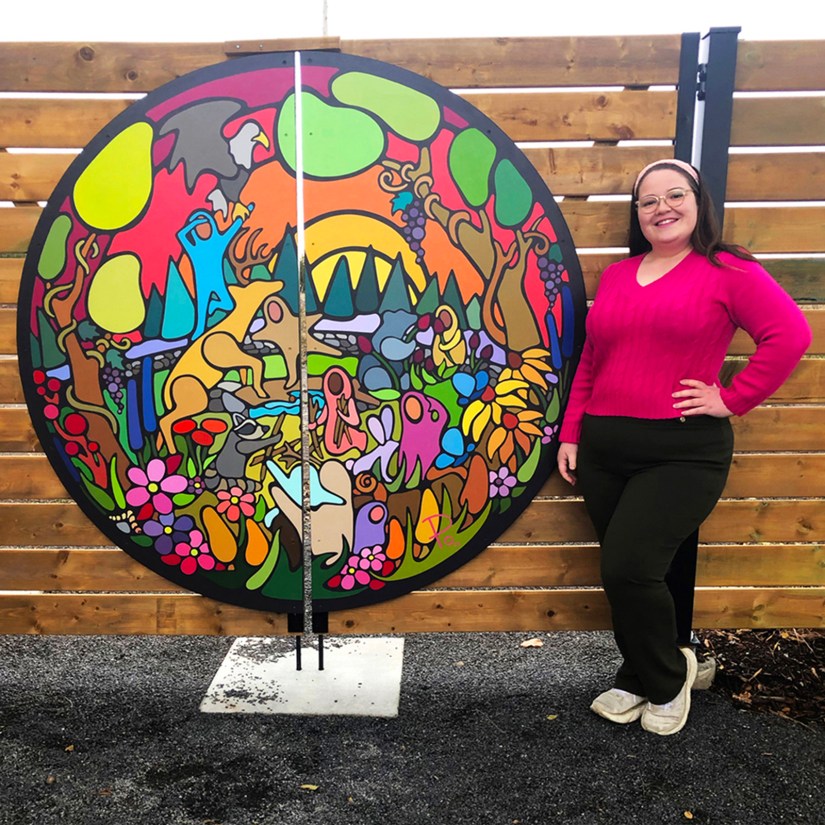 Kingston Artist Portia "Po" Chapman, wearing a vibrant pink sweater, stands smiling beside her circular "Exploring the Senses" mural on a wooden fence at Base 31 in the Sensory Garden during the spring of 2024.
