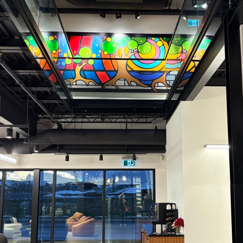 A first-floor mezzanine view of a wall-to-wall Storytelling Art mural by Portia "Po" Chapman, reflecting in glass railings to create an infinite visual illusion.