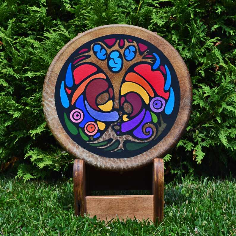 A gallery-quality painted rawhide frame drum by Kingston Artist Portia “Po” Chapman entitled "Sharing Wisdom," displayed on a wooden stand against a lush green cedar backdrop.
