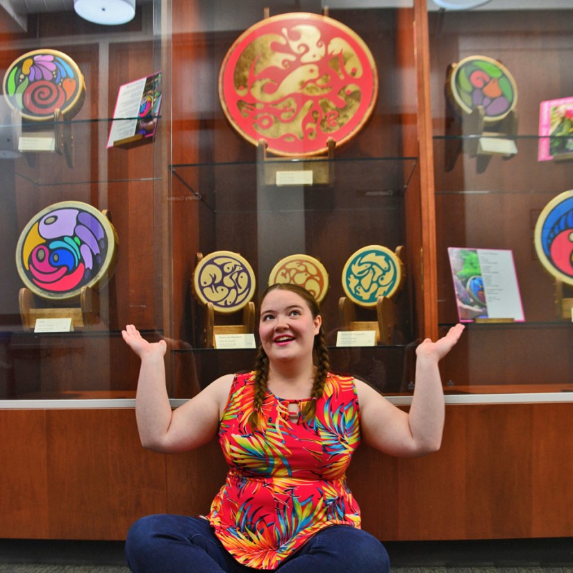 Kingston Artist Portia “Po” Chapman posing in front of her 15-piece "Drumming Sounds of Colour" exhibition display at the John M. Parrott Gallery, featuring multiple gallery-quality painted rawhide frame drums.