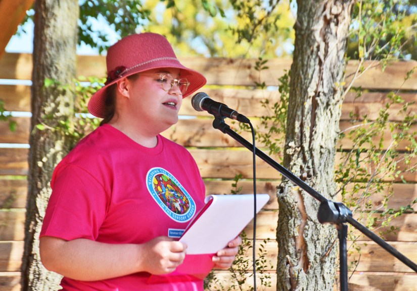 A portrait of Kingston artist Portia "Po" Chapman speaking into a microphone at the Sensory Garden grand opening, wearing a custom t-shirt featuring her "Exploring the Senses" freehand brush painted mural.