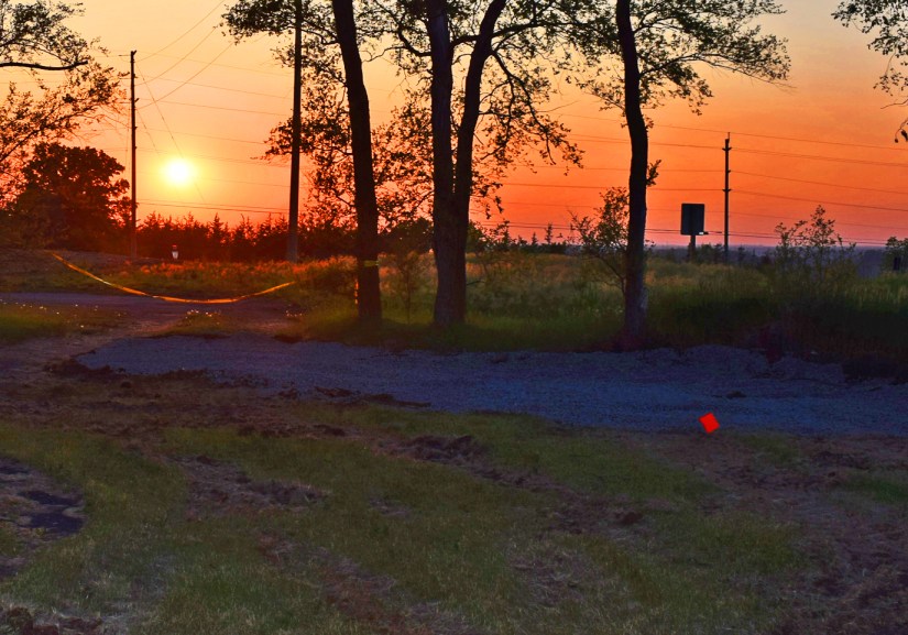 A vibrant orange and red sunset viewed through a silhouetted treeline at the Base31 construction site, with yellow caution tape marking the future "Exploring the Senses" mural location.