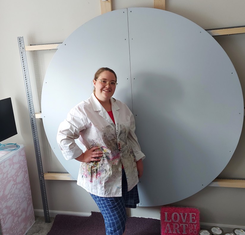 Kingston Mural Artist Portia Chapman in a paint-stained lab coat standing proudly in front of a giant, 5'-5" grey primed circular mural board mounted to a studio wall.