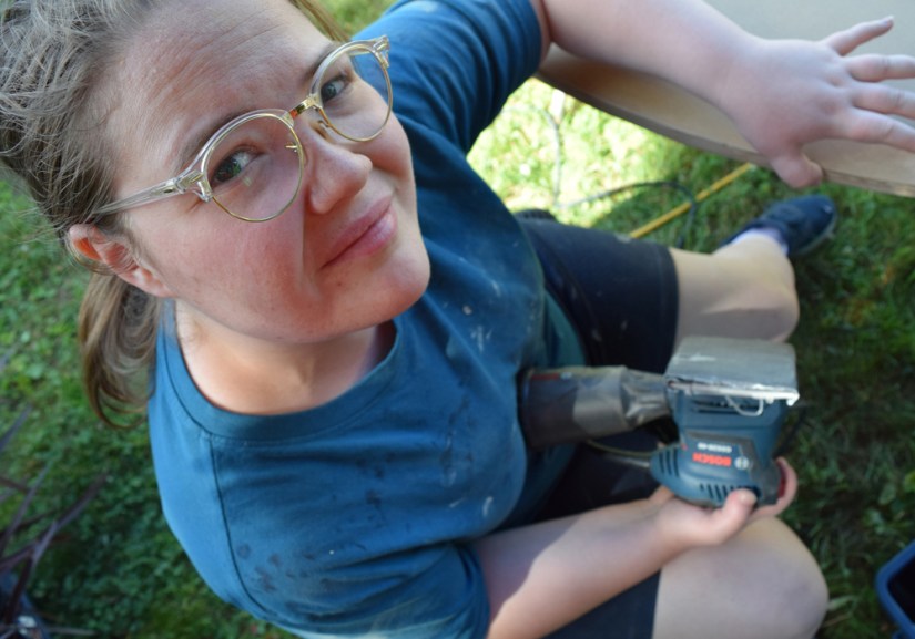Kingston Artist Portia Chapman working outside at her studio, using a Bosch power sander to smooth down the curved edge of a large wooden mural piece.