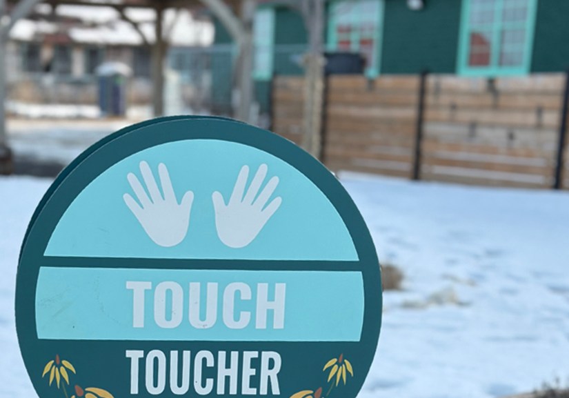 A bilingual "Touch / Toucher" sensory garden sign featuring white hand icons, colour-matched to the forest spruce green and seafoam blue of the adjacent historic WWII barracks building in winter.