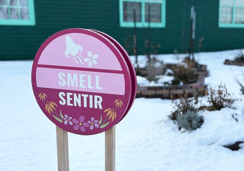 A circular bilingual "Smell / Sentir" sign in shades of cranberry and pink, featuring a nose and flower icon, set against a snowy garden and a forest green building.
