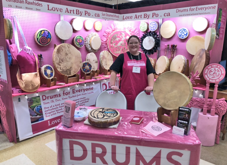 Kingston artist and frame drum expert maker Portia “Po” Chapman standing in her "Drums for Everyone" booth at The Maker’s Hand in Wellington, Ontario, showcasing a large, diverse collection of one-of-a-kind handcrafted rawhide frame drums that she made available for sale.