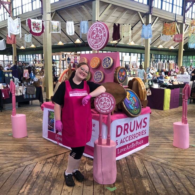 Kingston artist and drum making expert Portia “Po” Chapman standing at her vibrant "Drums for Everyone" interactive exhibition booth at Gather in the County's Textile Fair, featuring a variety of one-of-a-kind, handcrafted, and painted frame drums.