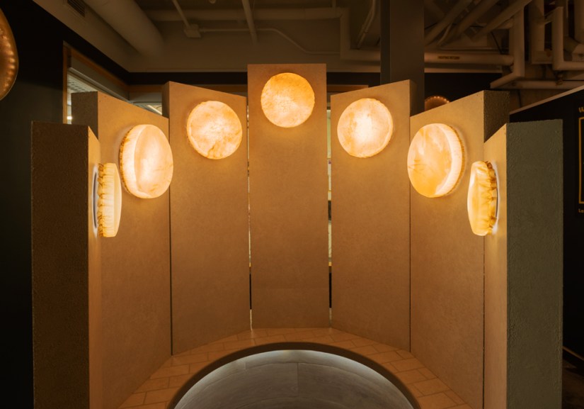 Interactive Glow Chamber installation featuring semi-circular partition walls mounted with circular, illuminated rawhide vessels casting a warm golden glow.