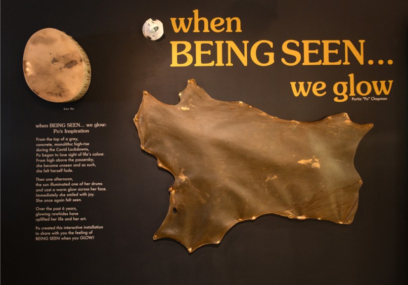 Exhibition title wall featuring large gold typography reading "when BEING SEEN... we glow" above a stretched translucent rawhide art piece and introductory text panel on a dark background.