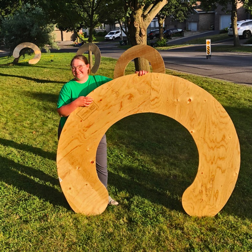 Kingston artist Portia Po Chapman standing outside her studio on the grass, smiling behind large cut plywood arc sections leaning against trees.