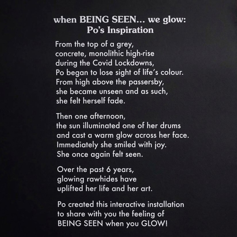 Close-up view of the white vinyl artist statement graphic on the dark title wall, titled "when BEING SEEN... we glow: Po's Inspiration," detailing Kingston artist Portia Po Chapman's story of finding light and joy through glowing rawhides.