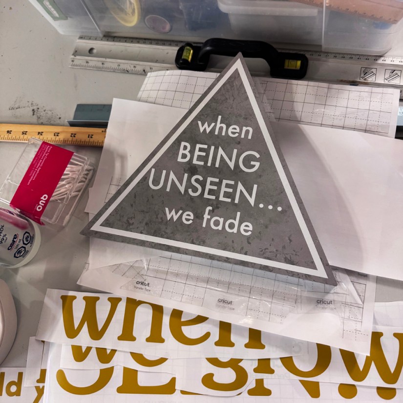 Overhead view of Kingston artist Portia Po Chapmanโs studio workbench displaying the triangular grey "when Being Unseen... we fade" caution sign alongside golden vinyl lettering on transfer paper during the creation of her interactive installation.
