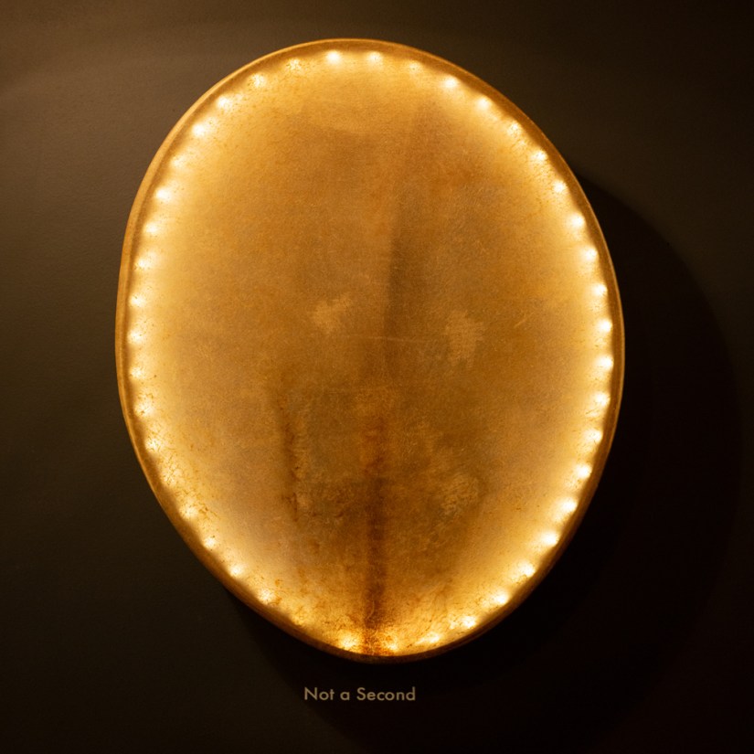 Oval bison rawhide drum titled Not a Second mounted on a dark gallery wall, backlit by circular LED points casting a warm marquee glow along its rim.