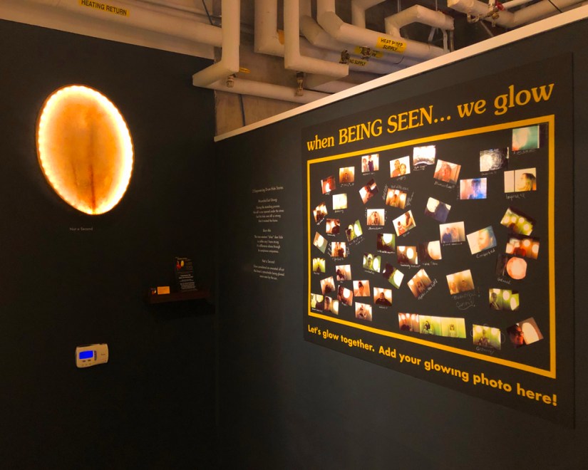 Dark gallery wall featuring a glowing bison rawhide drum mounted above a small shelf holding promotional business cards and rack cards beside an interactive selfie photo board.