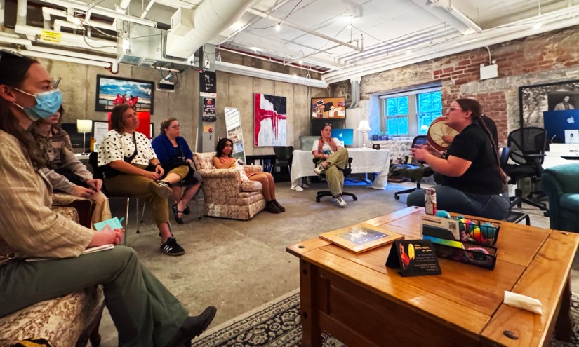 Kingston artist Portia Po Chapman presenting a rawhide drum during a private art talk at the Kingston Arts Council office with a promotional tent card featured on the foreground coffee table.
