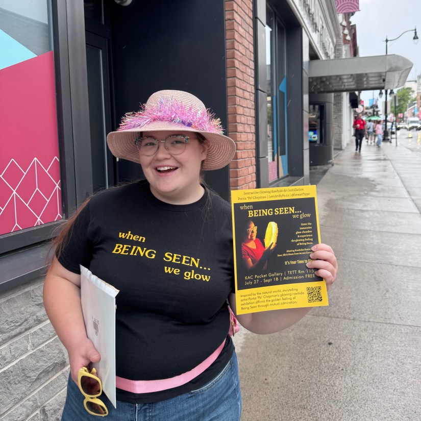 Kingston artist Portia Po Chapman wearing a pink tinsel hat and custom exhibition t-shirt while holding a Being Seen poster on a downtown Kingston street.