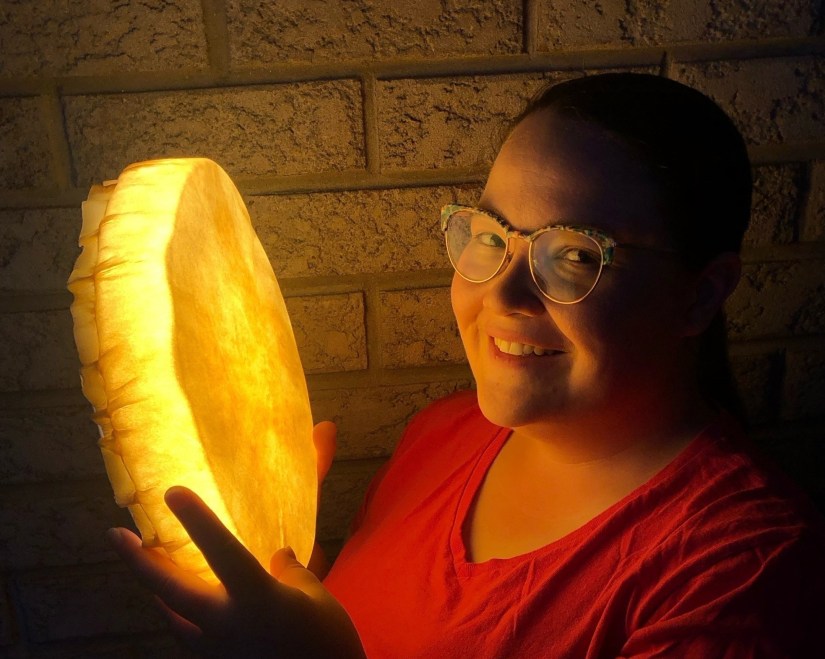 Kingston artist Portia Po Chapman holding a glowing illuminated rawhide frame drum at night outside a brick studio wall, radiating a warm golden light across her face and red shirt.