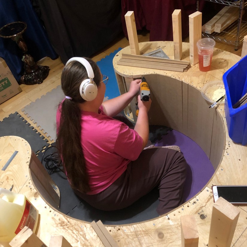 Kingston artist Portia Po Chapman seated inside the center of a curved wooden plinth using a pneumatic brad nailer to attach flexible sheathing to vertical support studs.
