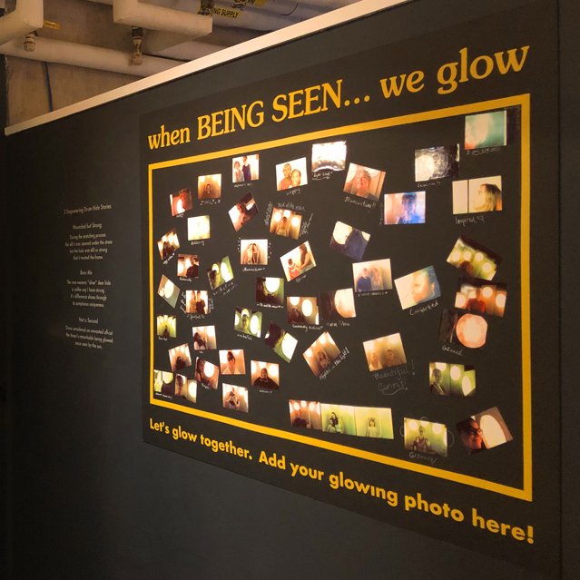 A dark gallery wall displaying a gold-framed interactive photo board titled "when BEING SEEN... we glow" covered in visitor selfie photos and handwritten notes, alongside custom wall text explaining three empowering drum hide stories by Kingston artist Portia Po Chapman.