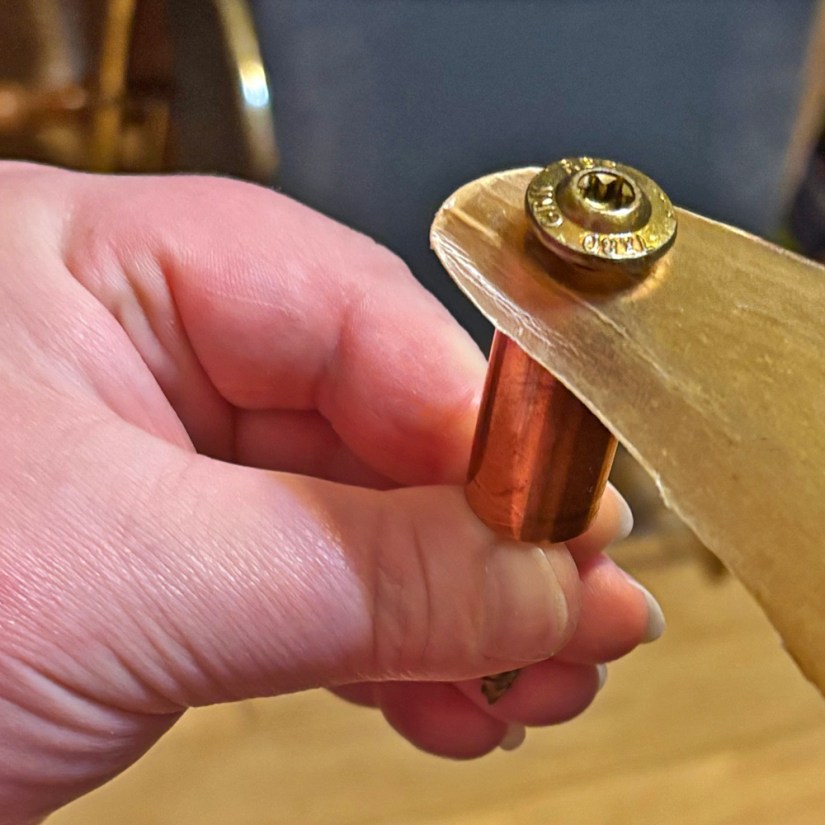 A close-up view of Portia Po Chapman's hand holding a trial assembly showing a brass star-headed screw passing through a drilled hole in a trimmed deer rawhide corner and a copper pipe spacer.
