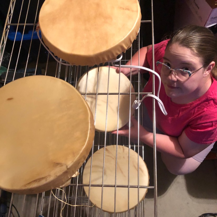 Top-down view of Kingston artist Portia Po Chapman in her woodshop, wearing glasses and a pink t-shirt, carefully placing an illuminated rawhide vessel onto a wire drying rack. Several other partially dried rawhide vessels are already sitting on the rack