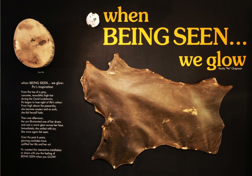 The completed black exhibition title wall for "when Being Seen... we glow" by Kingston artist Portia Po Chapman, featuring gold vinyl lettering, a full deer rawhide mounted 1.5 inches off the wall with copper pipe spacers, an artist statement, and an egg-shaped illuminated silver deer rawhide drum.