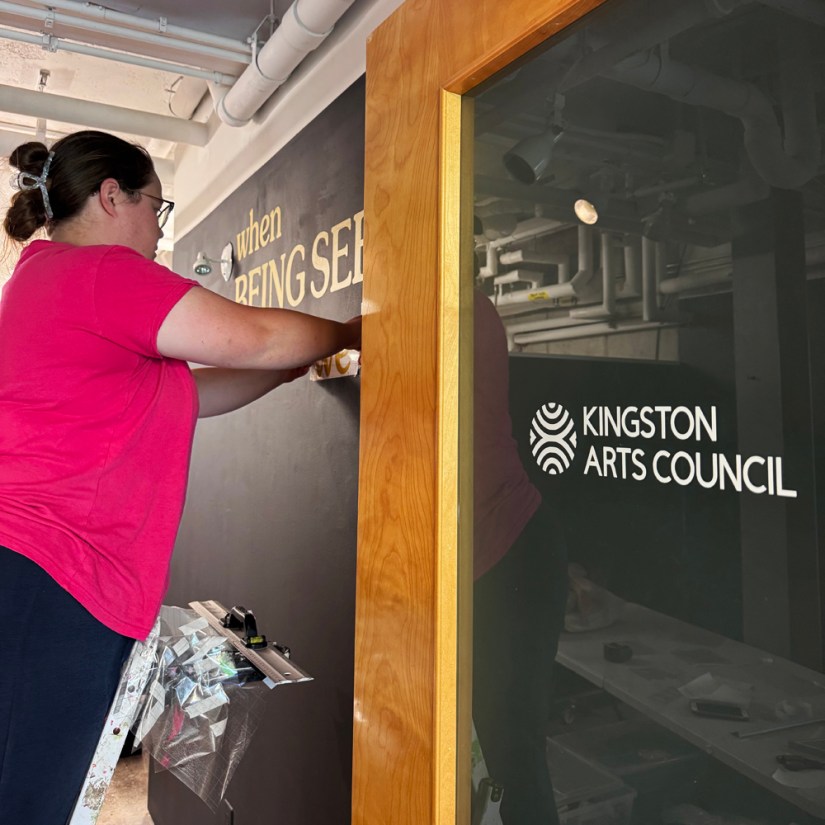 Side-angle view of Kingston artist Portia Po Chapman standing on a ladder inside the Pocket Gallery applying gold vinyl lettering reading "when BEING SEEN..." to the interior black title wall, with the Kingston Arts Council logo visible on the glass door frame in the foreground.