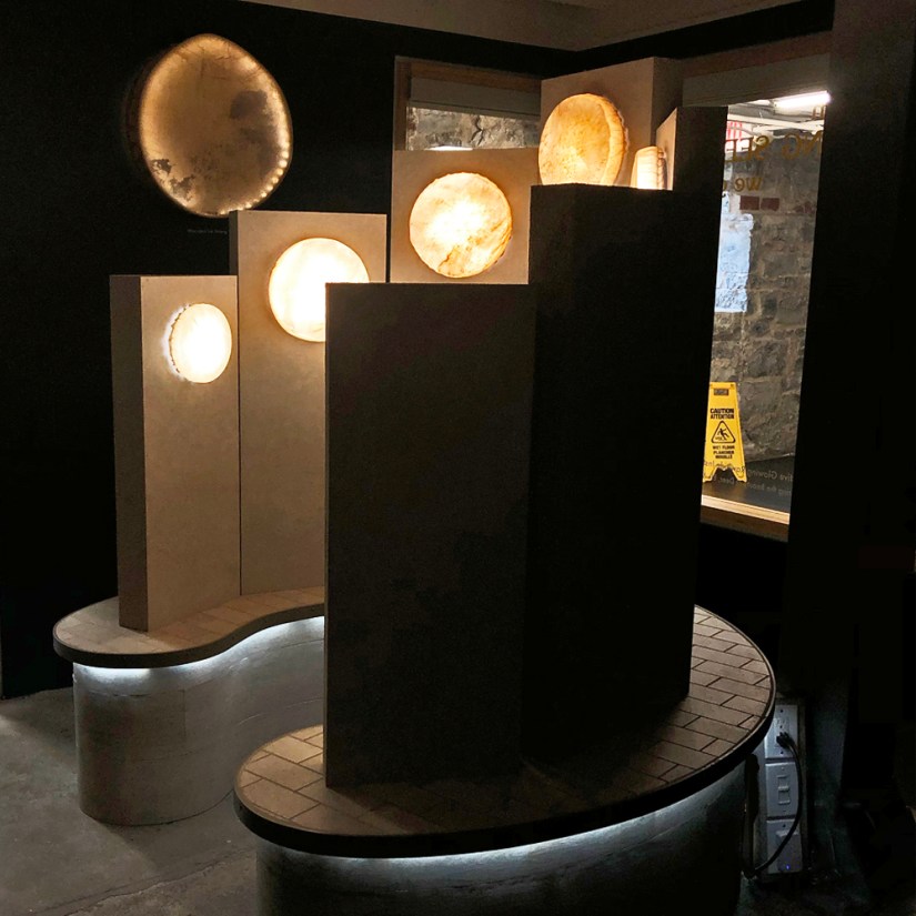 The illuminated Glow Chamber sculpture alongside a custom back-lit elk rawhide drum inside the Kingston Arts Council Pocket Gallery, framed by gallery windows, structural pillar, and entrance.